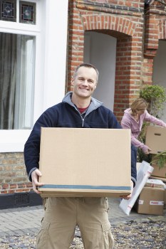 A man with a friendly expression, wearing a navy blue jacket and khaki trousers, is standing outside a residential brick house holding a large, sealed cardboard box with both hands. Behind him, a woman with blonde hair, dressed in a purple top, is seen bending down and carrying another box, with additional cardboard boxes around her, some open and filled with household items. The scene takes place on a cobbled street or driveway in a suburban area, with the house featuring white-framed windows and a small potted plant near the entrance. The overall lighting suggests daytime with natural light, and the environment appears to be a typical house relocation setting, illustrating a house removal or moving process, consistent with the services of Stockwell Man with Van in London.