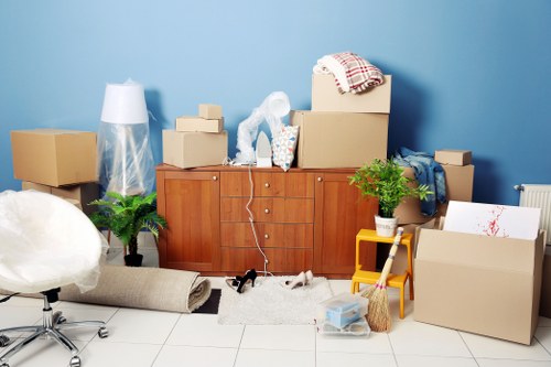The image depicts a room with a light blue wall and a white tiled floor, filled with various household items and packing materials. In the background, there is a wooden sideboard with multiple drawers, on top of which are several cardboard boxes, some sealed and others open, containing packing paper and miscellaneous belongings. A clear plastic bag filled with supplies and a white lamp are also positioned on the sideboard. To the left of the sideboard, a potted green fern plant is visible, along with a bundle of rolled-up carpets or rugs lying on the floor in front of it. On the right side, a small yellow step stool and a potted plant are present next to larger cardboard boxes, some partially open, and a white cardboard box with red paint splatters. Additionally, there are packing items such as bubble wrap, a broom, and a plastic storage container nearby. The scene suggests an active packing or moving process typical of house removals in the Stockwell area of London, indicated by the postcode inferred from the page context.